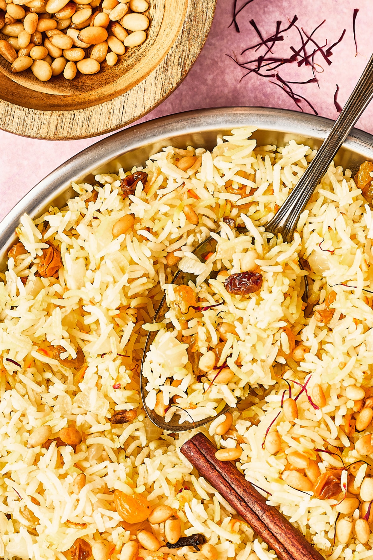 Overhead view of a silver serving bowl of yellow saffron rice garnished with golden raisins, pine nuts, a cinnamon stick and saffron strands. A small wooden bowl of toasted pine nuts sits to the side of the rice, and a serving spoon is in the bowl of rice.