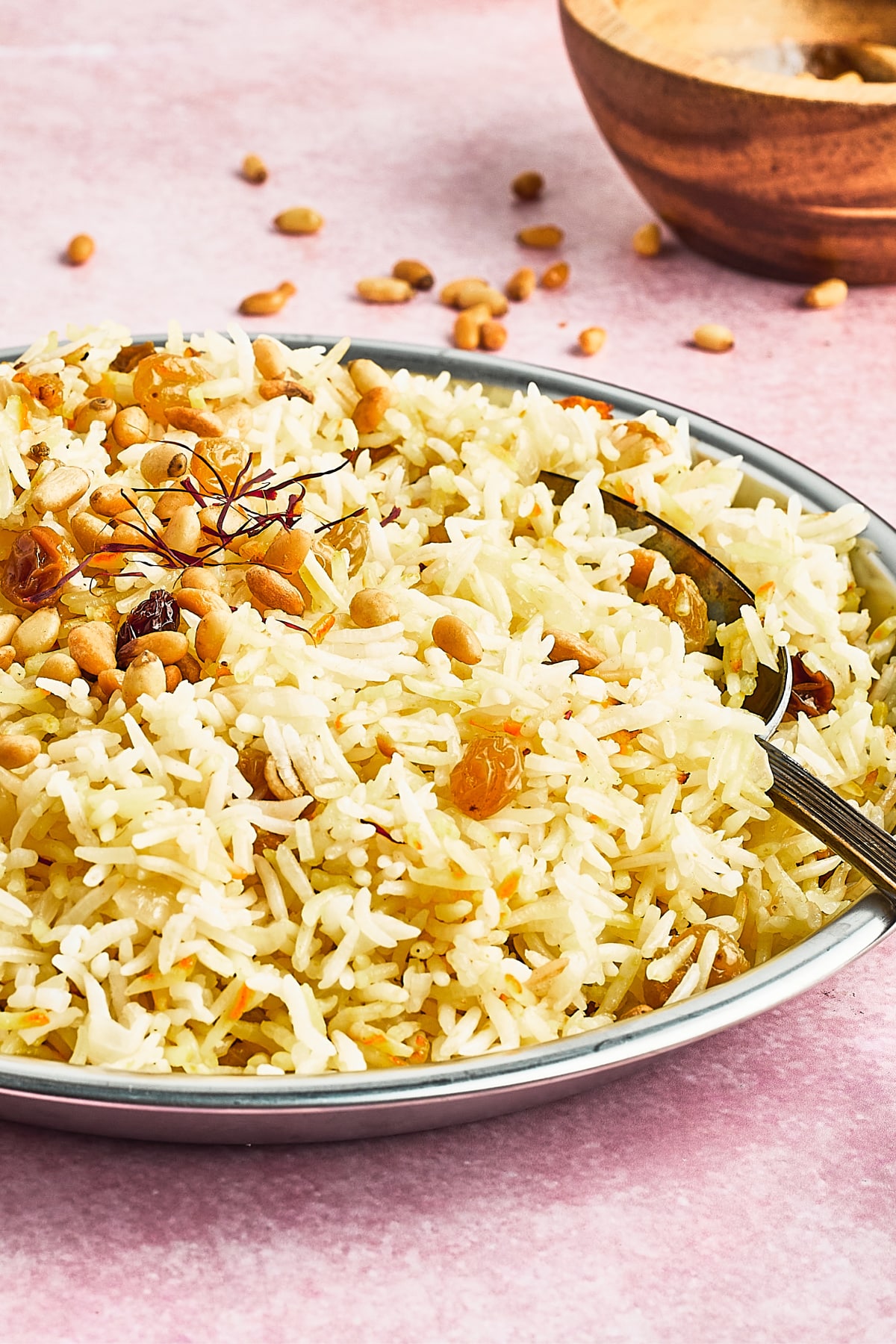 A shallow serving bowl of yellow saffron rice garnished with golden raisins, pine nuts, a cinnamon stick and saffron strands. A small wooden bowl of toasted pine nuts sits to the side of the rice, with pine nuts scattered on the pink surface.