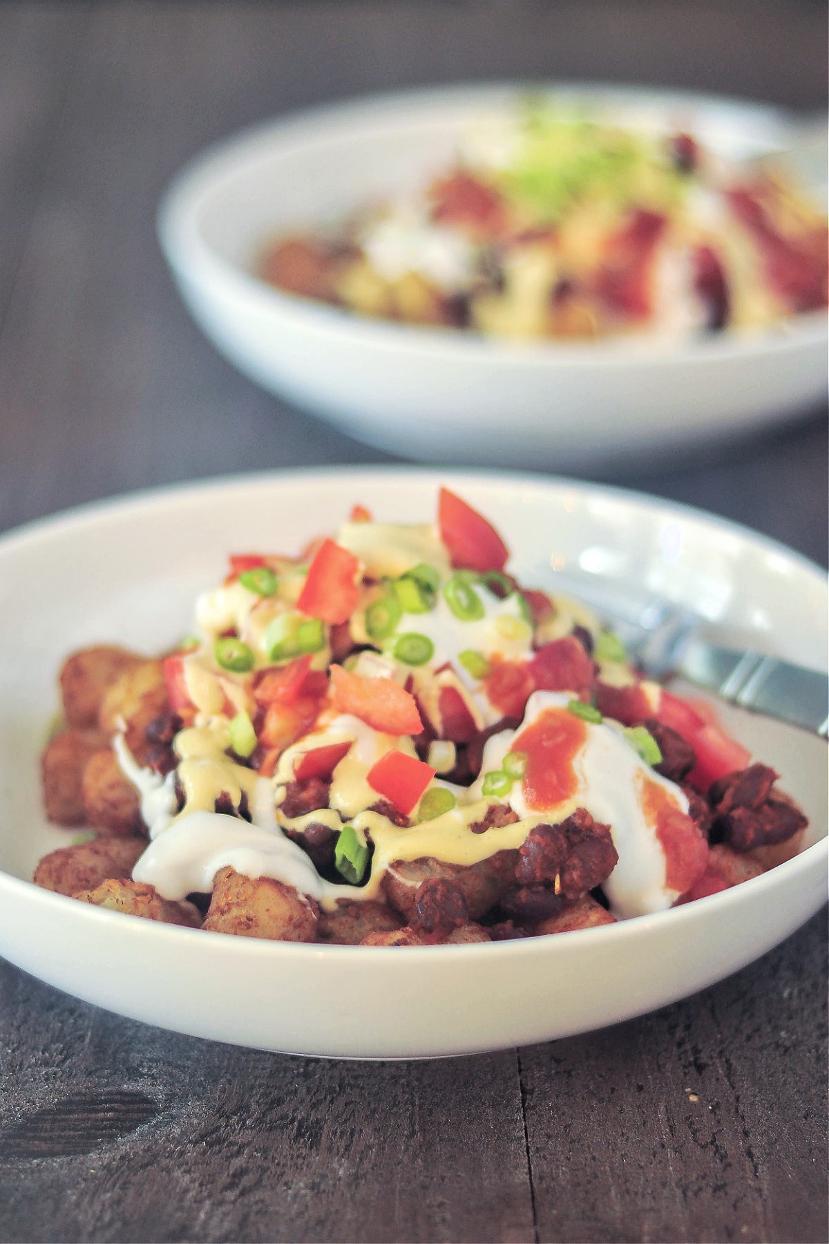 Two shallow white bowls filled with black bean tater tot nachos. Totchos are topped with garlic lemon sauce, tomatoes, jalapeños, salsa, and green onions.