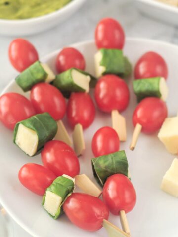 A round white plate with several caprese skewers sits on a marble table with other appetizers. A caprese skewer is two cherry tomatoes and a piece of cheese wrapped in a fresh basil leaf on a cocktail pick.