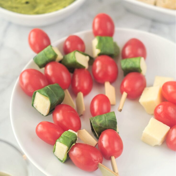 A round white plate with several caprese skewers sits on a marble table with other appetizers. A caprese skewer is two cherry tomatoes and a piece of cheese wrapped in a fresh basil leaf on a cocktail pick.
