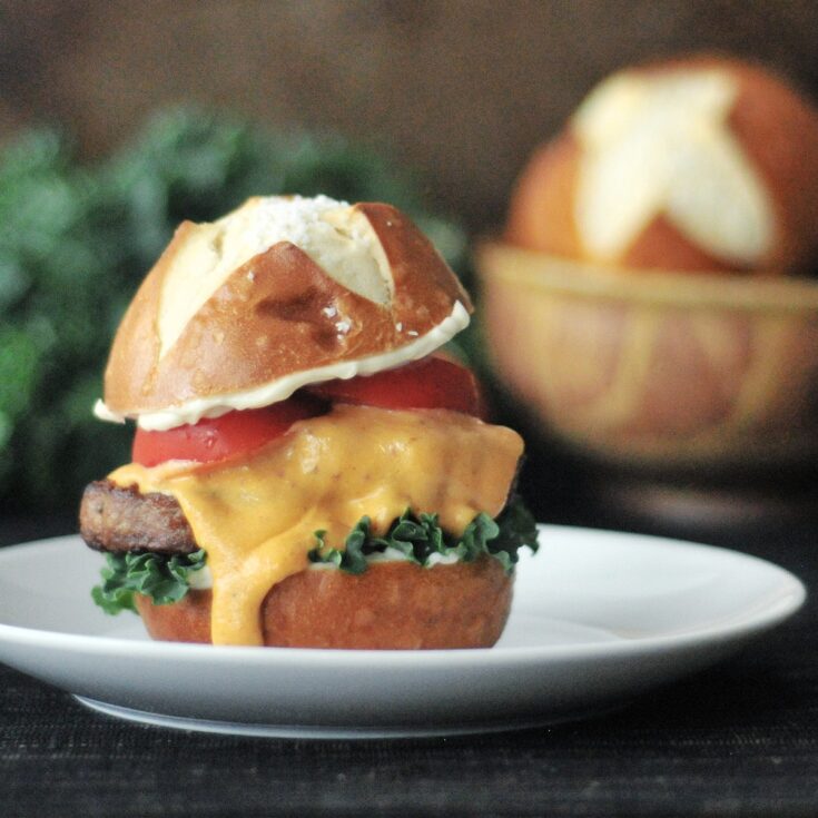 A pretzel bun veggie burger slider sits on a white plate. The slider has kale, tomato slices, and a vegan cheese sauce dripping down the side. A large bunch of kale, one whole tomato, and a bowl of pretzel buns are blurred in the background.