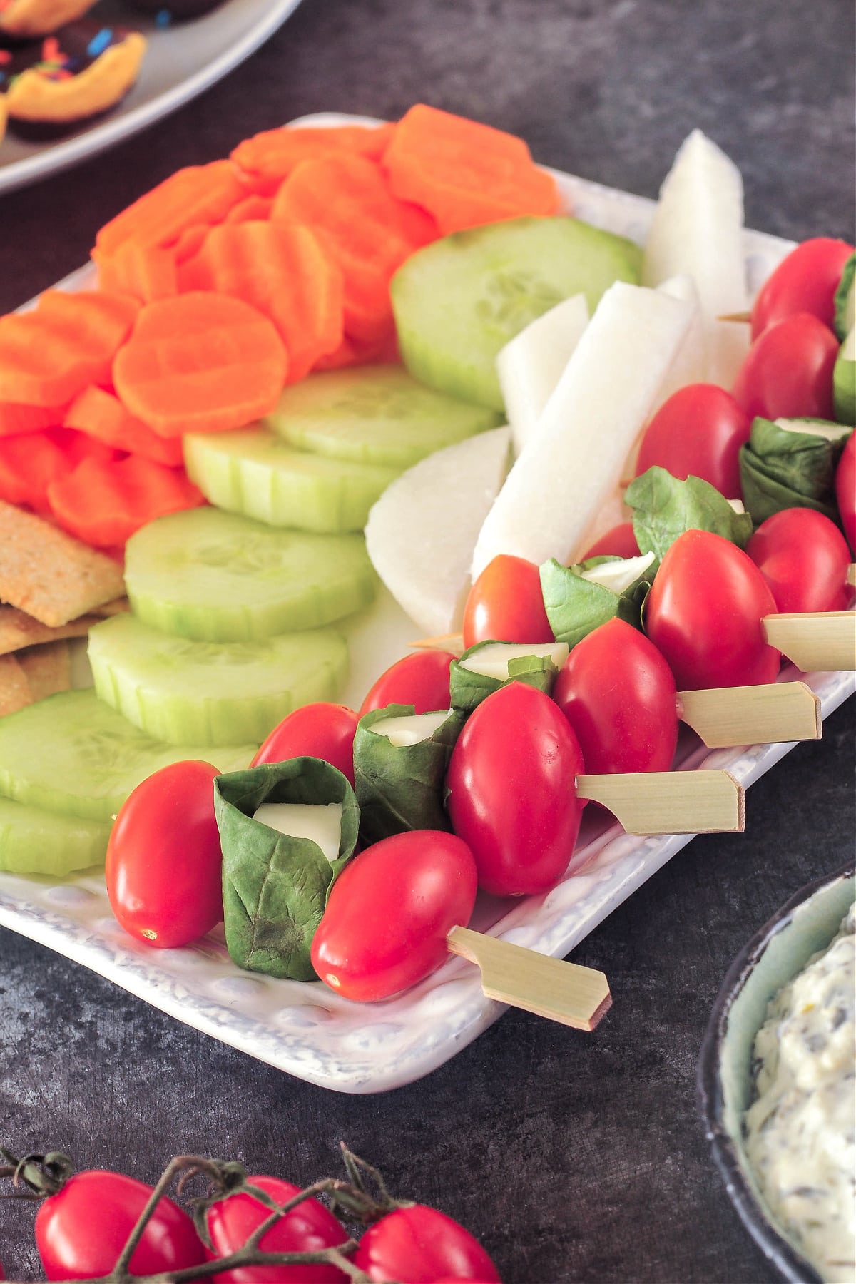 Several caprese skewers are lined up on a square white platter that also holds crackers and slices of jicama, cucumber, and carrot. A caprese skewer is two cherry tomatoes and a piece of cheese wrapped in a fresh basil leaf on a cocktail pick.