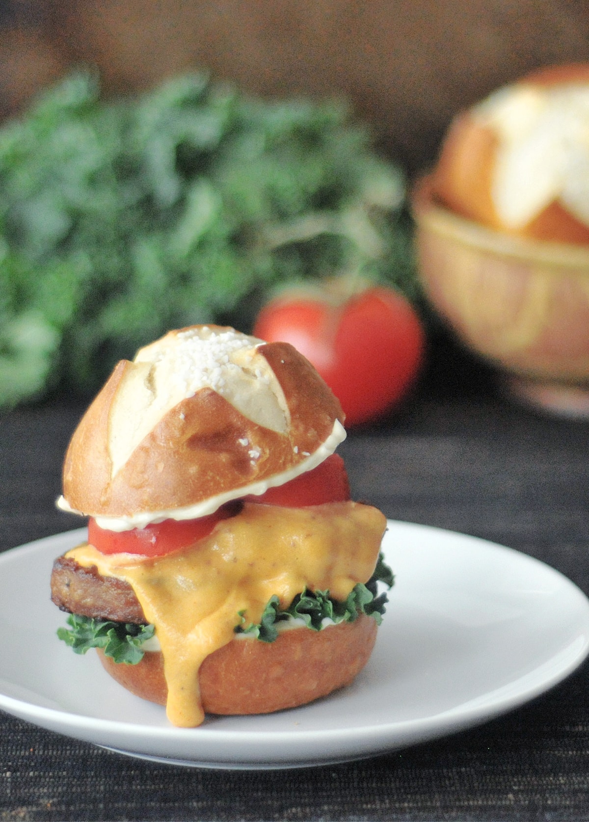A pretzel bun veggie burger slider sits on a white plate. The slider has kale, tomato slices, and a vegan cheese sauce dripping down the side. A large bunch of kale, one whole tomato, and a bowl of pretzel buns are blurred in the background.