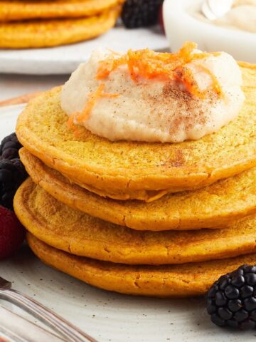 Close up view of a stack of four bright orange carrot pancakes sits on a white plate with blackberries and raspberries, and a fork and knife. Pancakes are topped with a maple cashew cream and grated carrot.