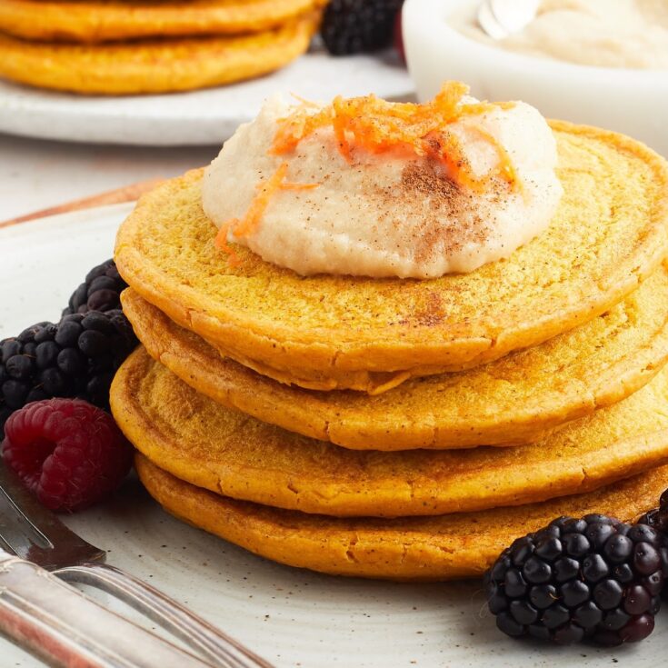 Close up view of a stack of four bright orange carrot pancakes sits on a white plate with blackberries and raspberries, and a fork and knife. Pancakes are topped with a maple cashew cream and grated carrot.