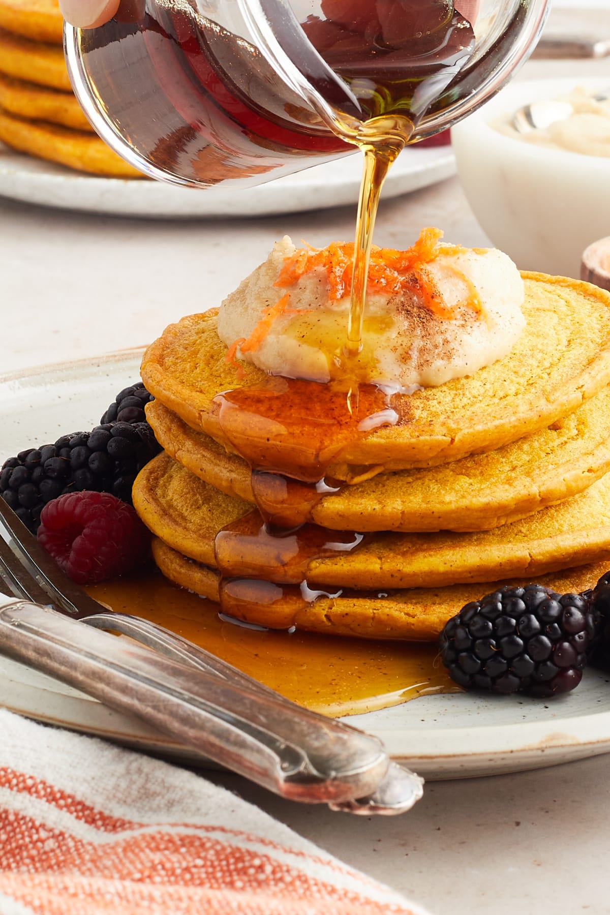 Maple syrup is being poured over a stack of four bright orange carrot pancakes topped with a maple cashew cream and grated carrot. Berries and a fork and knife sit on the plate with the pancakes.