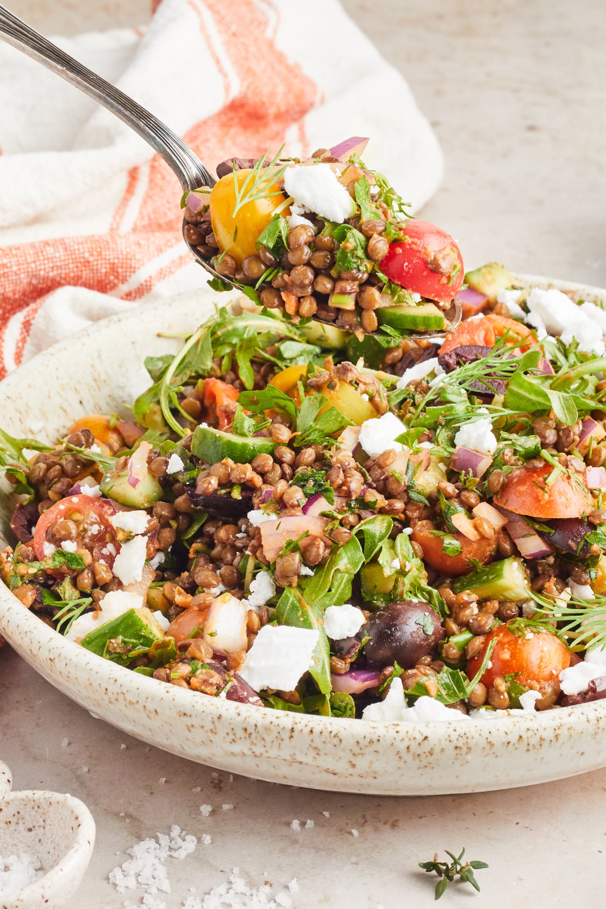 A spoon of salad hovers over a serving bowl of colorful Greek salad with lentils, sliced cherry tomatoes, sliced cucumber, fresh herbs, dairy free feta cheese. A small bowl of bright orange sun dried tomato dressing sits on the side with an orange and white striped cloth napkin.