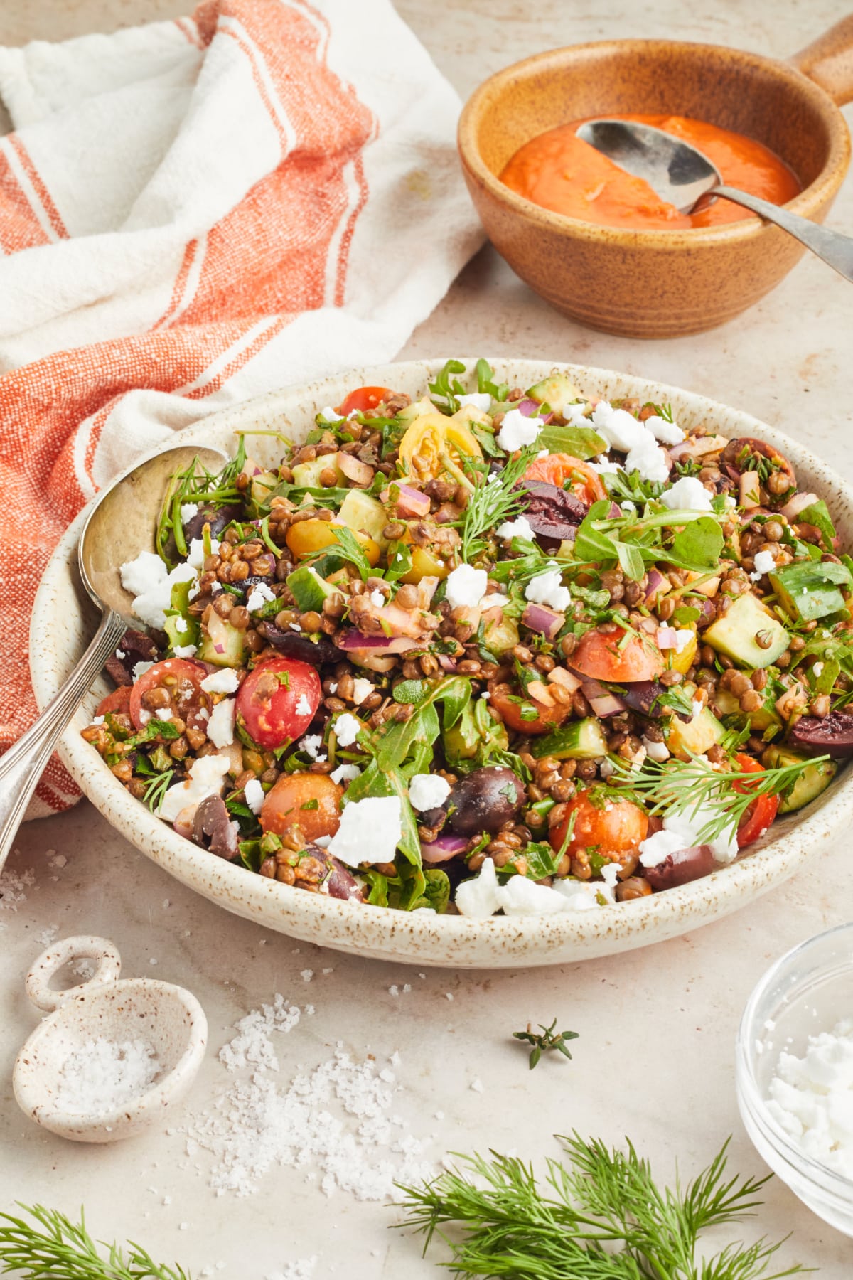 Serving bowl of colorful Mediterranean lentil salad consisting of lentils, sliced cherry tomatoes, sliced cucumber, fresh herbs, dairy free feta cheese. A small bowl of bright orange sun dried tomato dressing sits on the side with an orange and white striped cloth napkin.