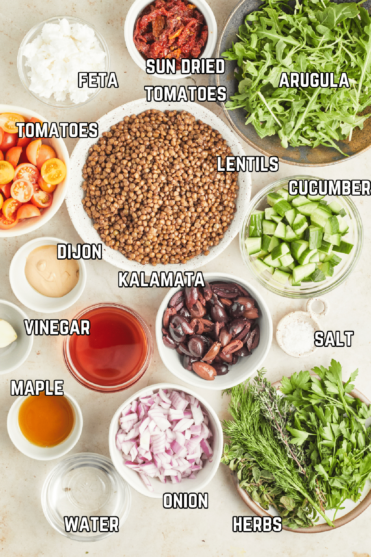 Overhead view of ingredient to make Mediterranean salad with lentils: crumbled feta, sun dried tomatoes, arugula, lentils, cucumber, tomatoes, Dijon, vinegar, kalamata olives, maple syrup, onion, fresh herbs, and water.