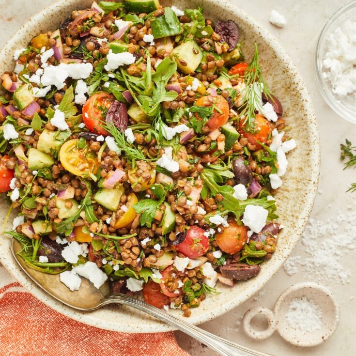 Overhead view of a serving bowl of colorful Greek lentil salad consisting of lentils, sliced cherry tomatoes, sliced cucumber, fresh herbs, dairy free feta cheese.