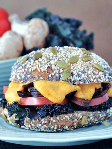 A mushroom melt sandwich with curly kale, tomato slices, mushrooms and cheese sauce on a seeded bun.