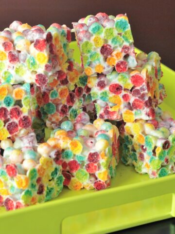 Squares of rainbow colored crispy cereal treats are stacked in a bright green serving tray. Treats are made from Trix cereal and marshmallows.