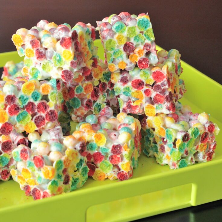 Squares of rainbow colored crispy cereal treats are stacked in a bright green serving tray. Treats are made from Trix cereal and marshmallows.