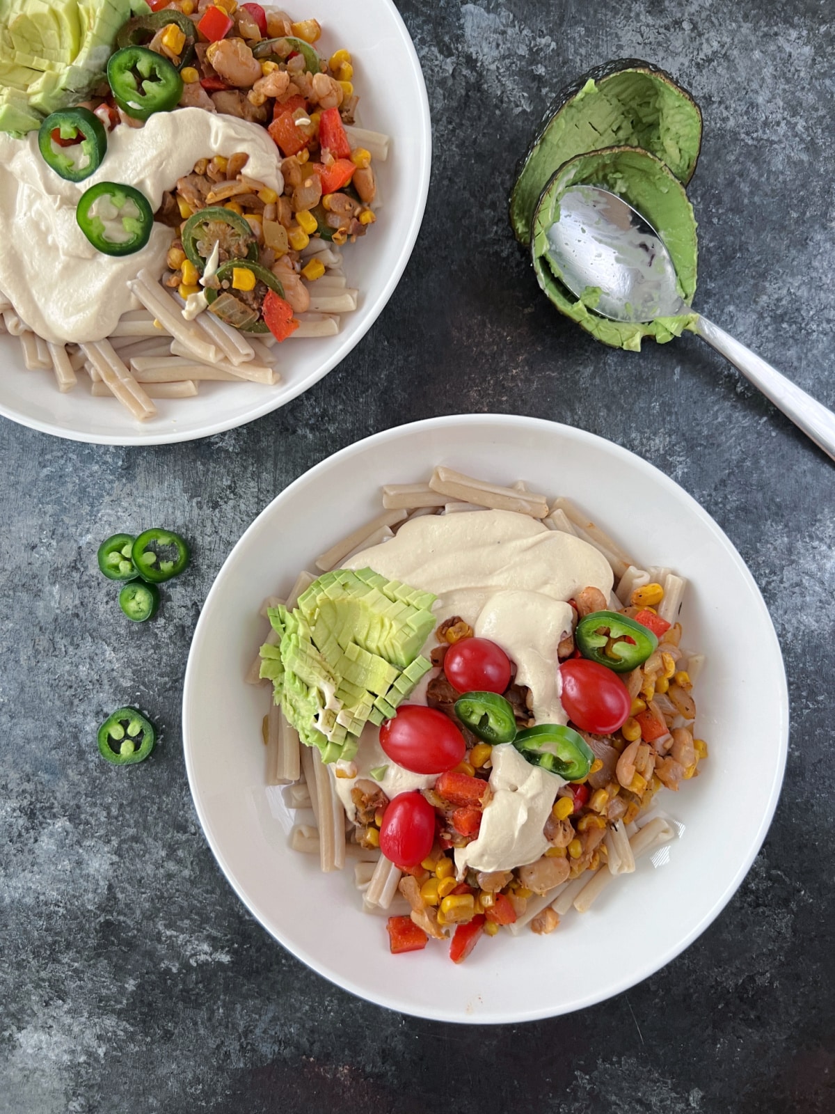 Overhead view of two white shallow bowls of Tex Mex pasta. Pasta has cheese sauce, beans, corn, three kinds of peppers, southwest spices, and is garnished with cherry tomatoes, jalapeno slices, and avocado.