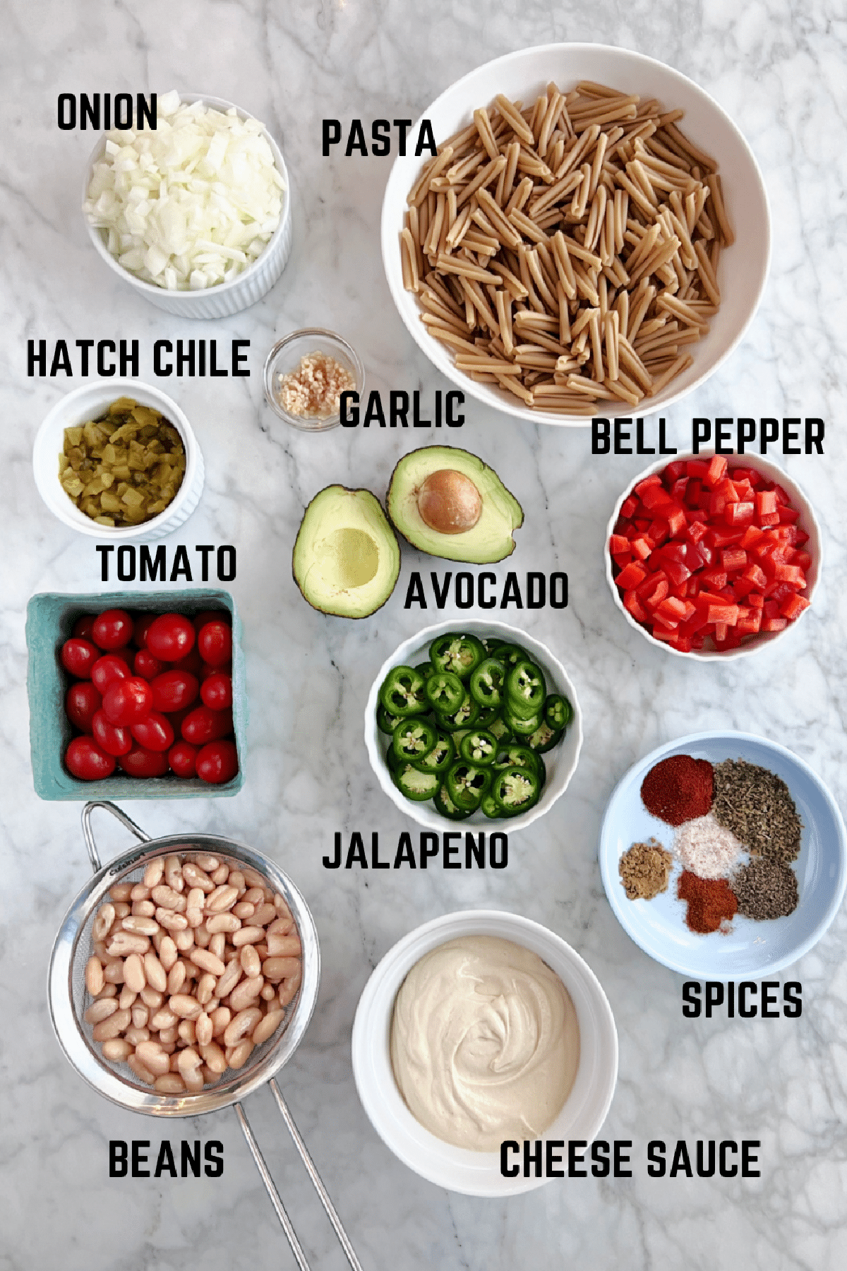 Overhead view of ingredients to make spicy southwest mac and cheese: pasta, onion, garlic, Hatch chiles, tomato, jalapeno, bell pepper, avocado, beans, cheese sauce, and southwest spices.