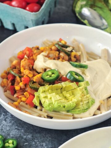 Close up of a white shallow bowl of Tex Mex pasta. Pasta has beans, corn, three kinds of peppers, southwest spices, and is garnished with cherry tomatoes, jalapeno slices, and avocado.
