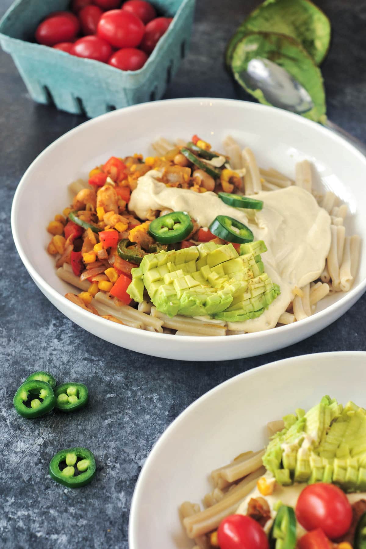 Close up of a white shallow bowl of Southwest pasta. Pasta has beans, corn, three kinds of peppers, southwest spices, and is garnished with cherry tomatoes, jalapeno slices, and avocado.