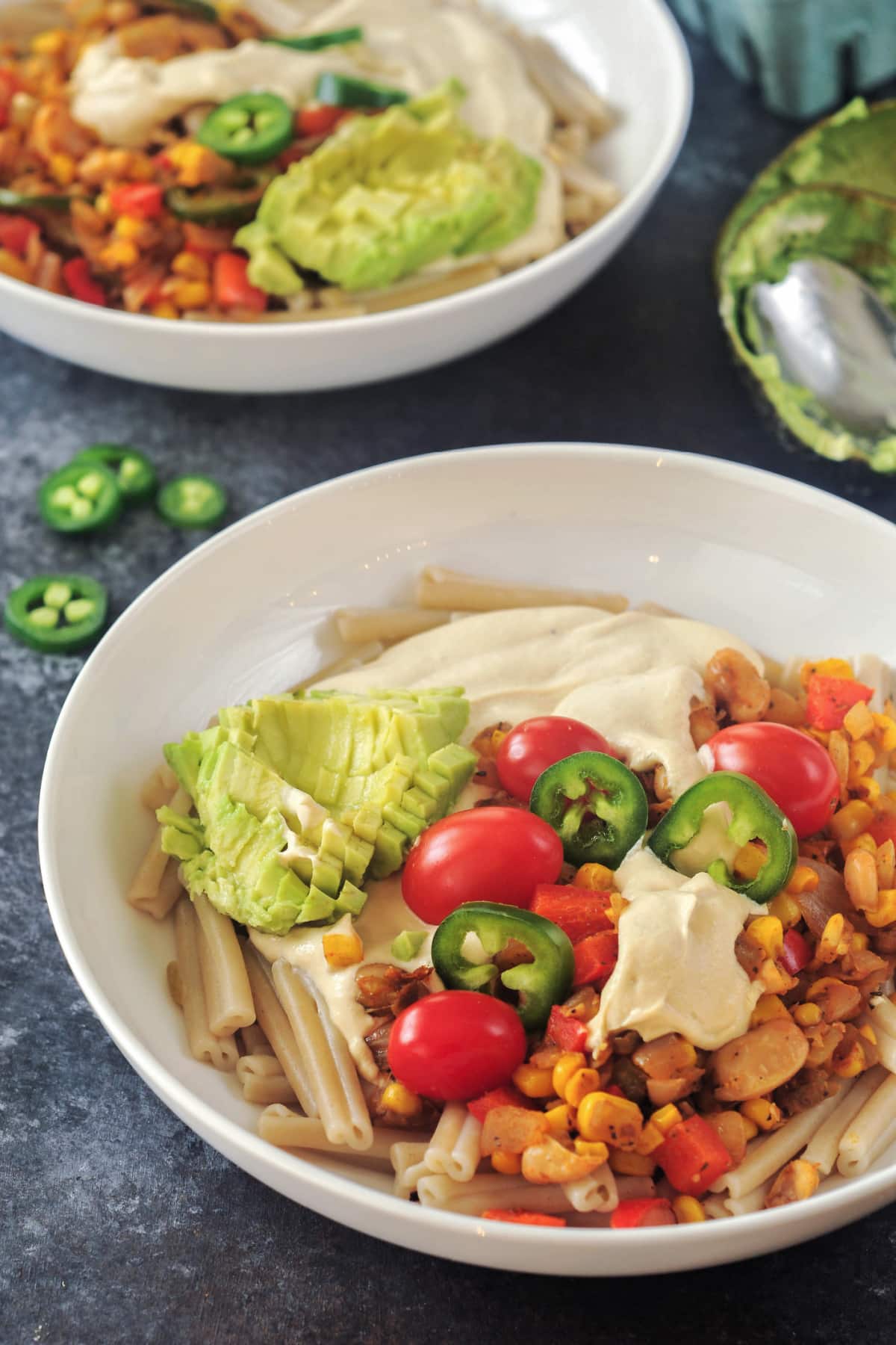 Two white shallow bowls of Southwest Mac and Cheese. Pasta has cheese sauce, beans, corn, three kinds of peppers, southwest spices, and is garnished with cherry tomatoes, jalapeno slices, and avocado.