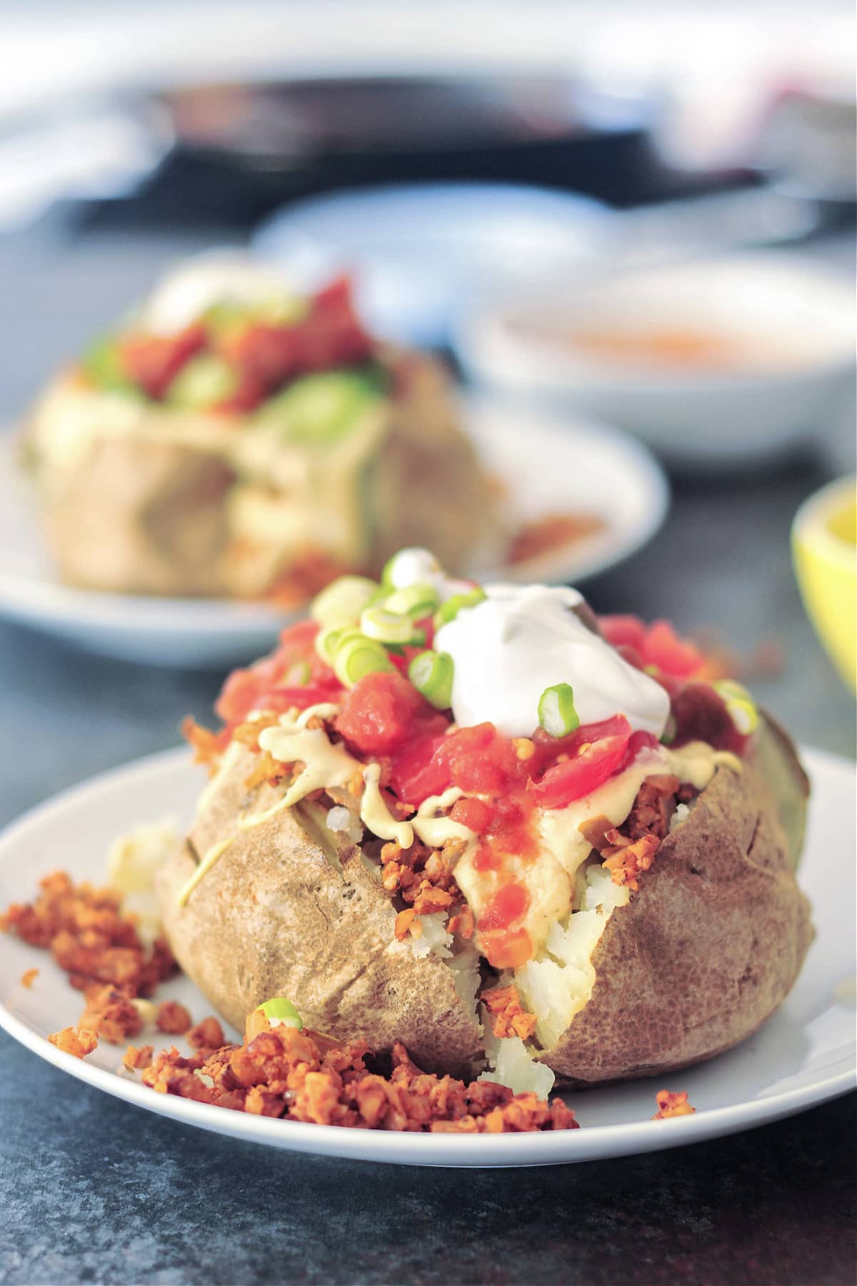 two taco stuffed baked potatoes served on a white plate; potatoes are stuffed with taco meat, cheese sauce, salsa, sliced jalapeños, sour cream and sliced green onion. Small bowls of salsa, cheese sauce, chopped tomatoes and green onions sit on the side.