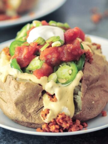 A taco stuffed baked potato served on a white plate; potato is stuffed with taco meat, cheese sauce, salsa, sliced jalapeños, sour cream and sliced green onion.