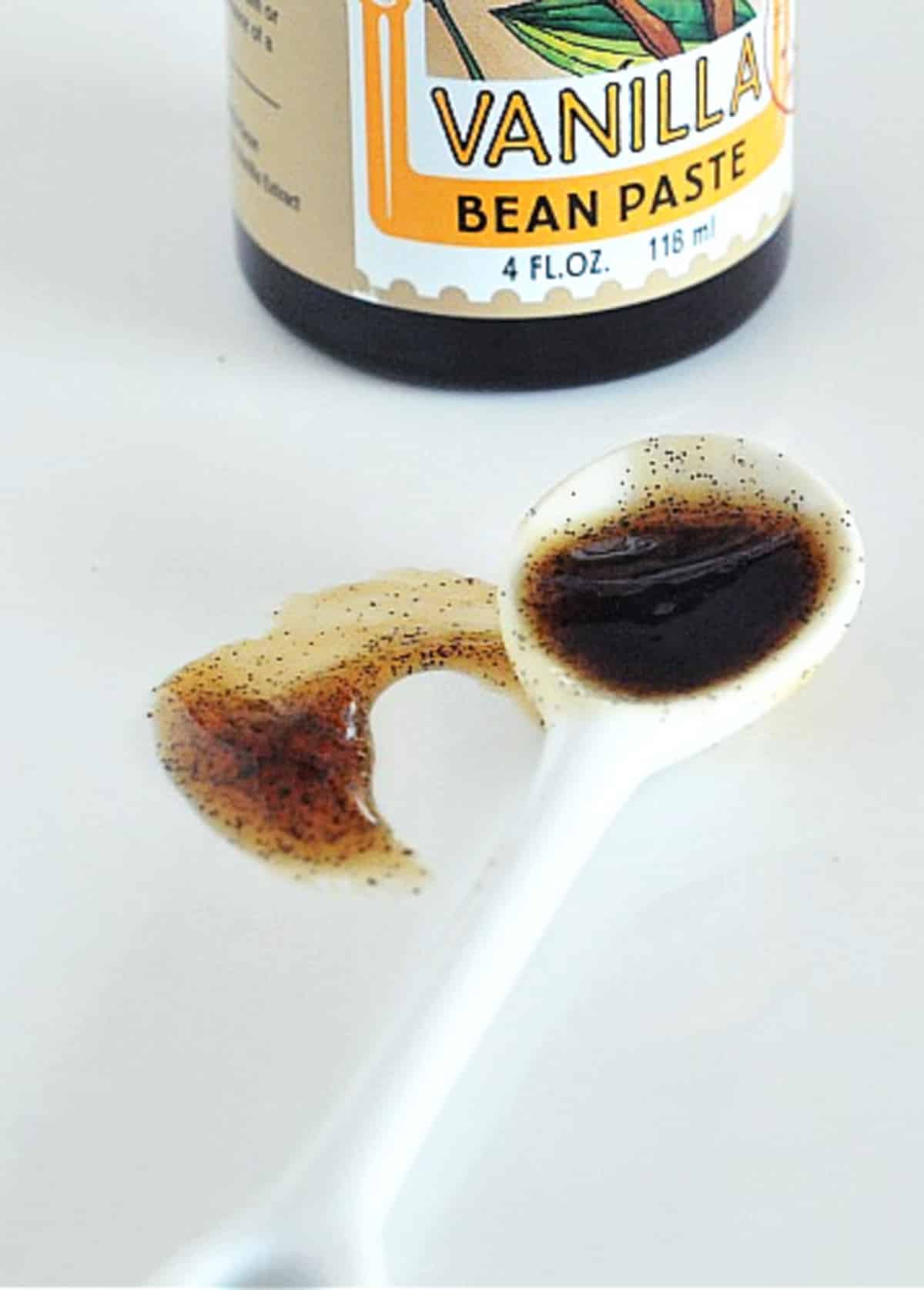 A white ceramic spoon of vanilla bean paste with a smear of the paste on a shiny white surface next to the spoon. The vanilla paste jar sits behind the spoon, only partially in frame of the photo.