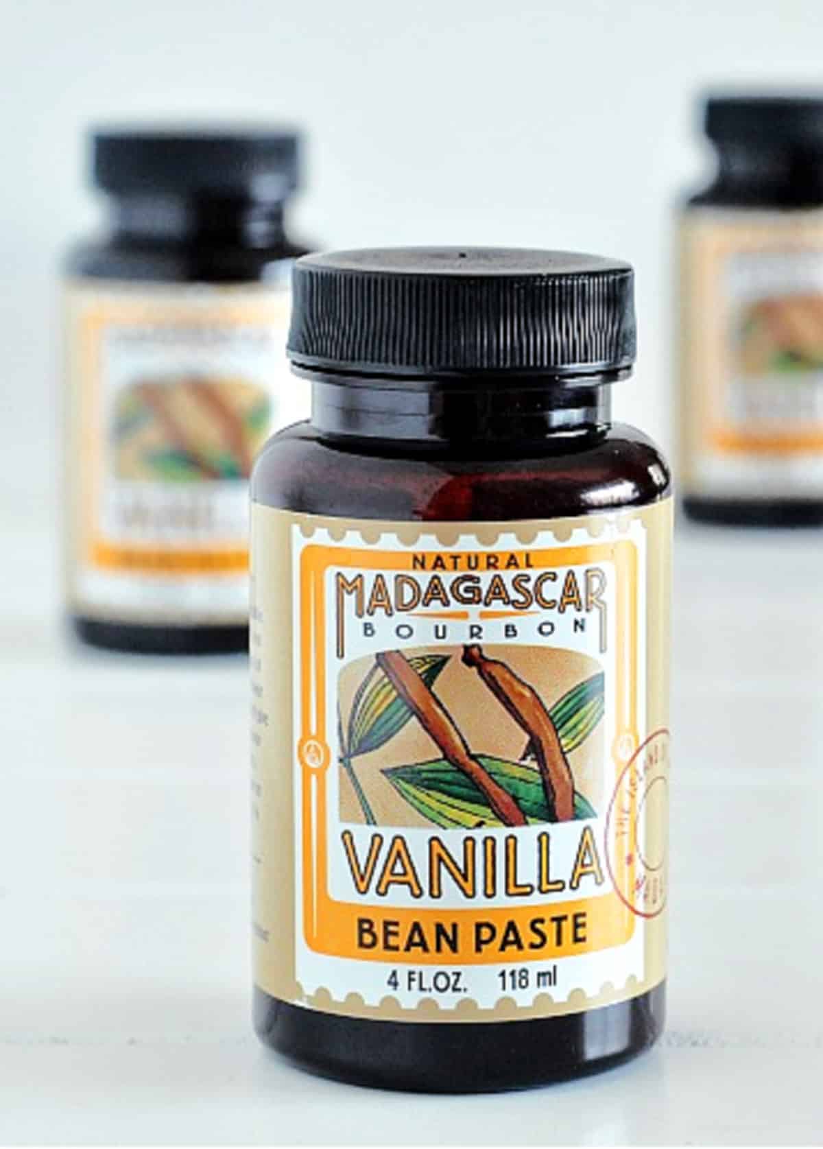 Three jars of vanilla bean paste sit on a white surface; two of the jars are blurred in the background.