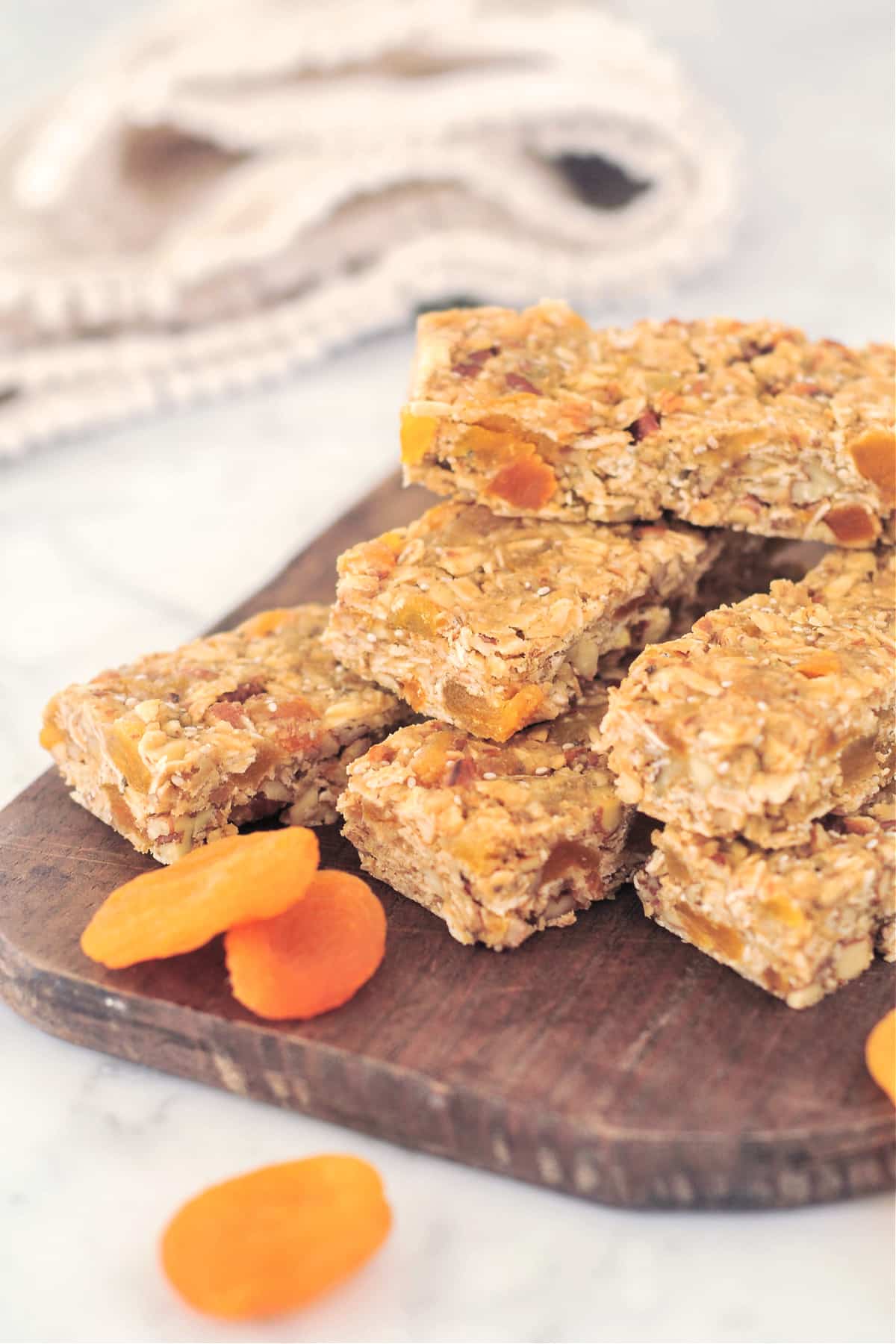Several apricot almond granola bars are stacked on a dark wooden board, with two dried apricots.