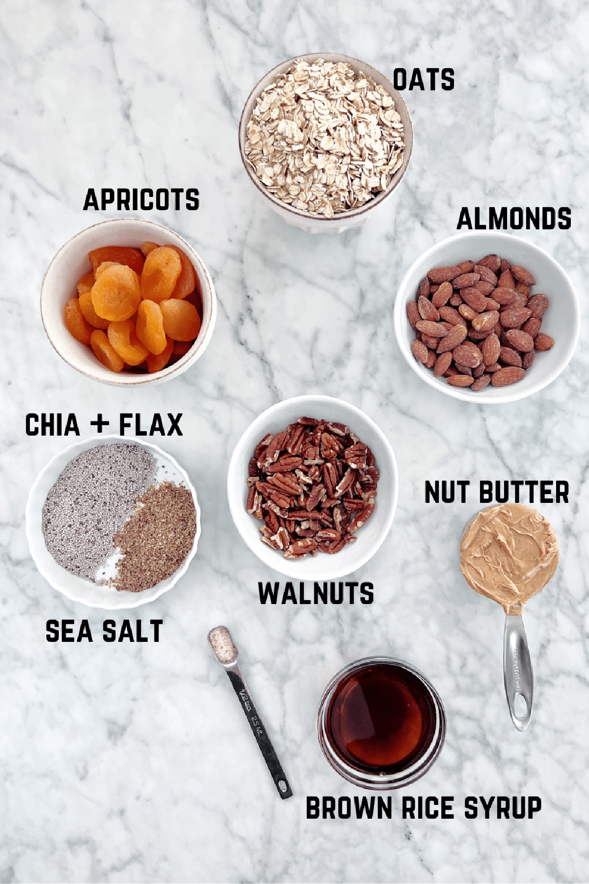 Overhead view of ingredients to make granola bars: oats, dried apricots, almonds, walnuts, nut butter, brown rice syrup, salt, chia, and flax.