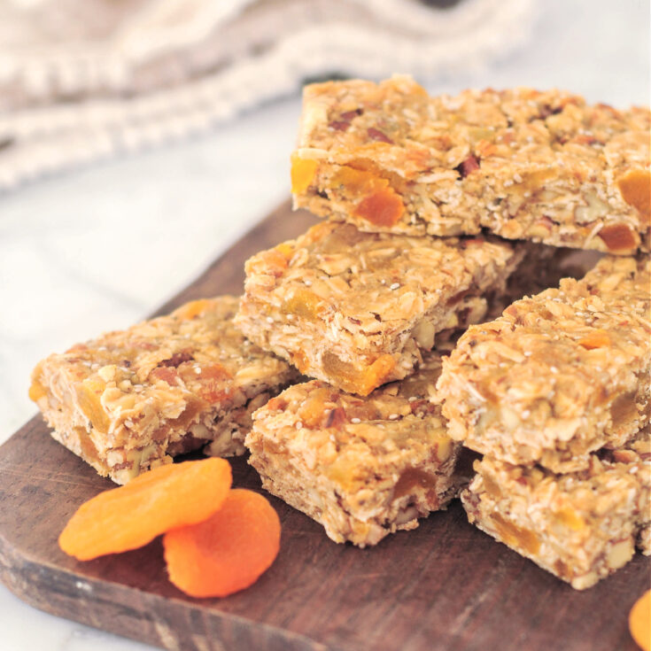 Several apricot granola bars are stacked on a dark wooden board, with two dried apricots.