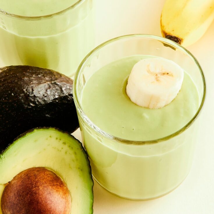 Two glasses of light green avocado drink garnished with banana slices. Whole bananas and an avocado sliced in half sit on the side.