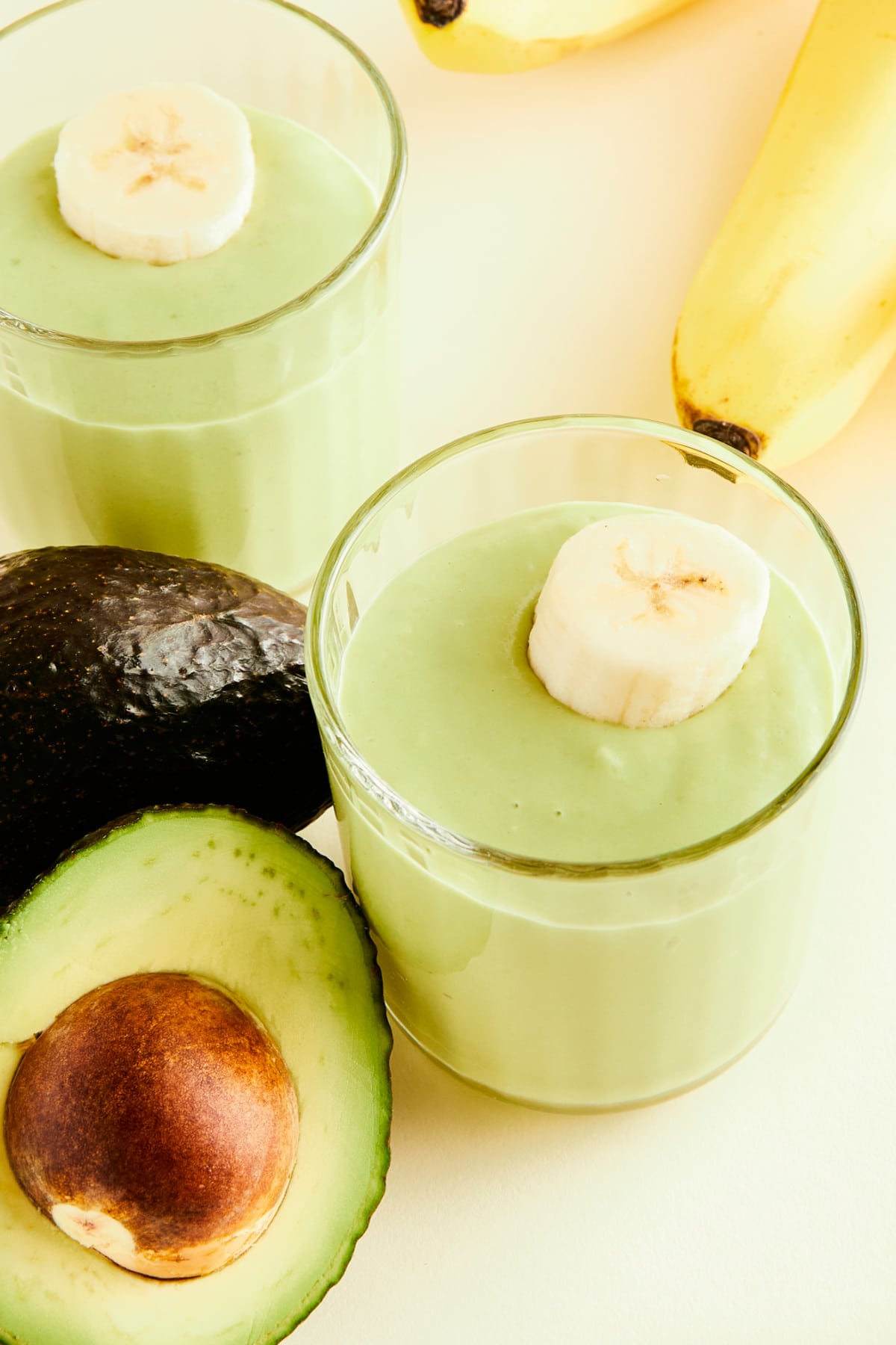 Two glasses of light green avocado drink garnished with banana slices. Whole bananas and an avocado sliced in half sit on the side.