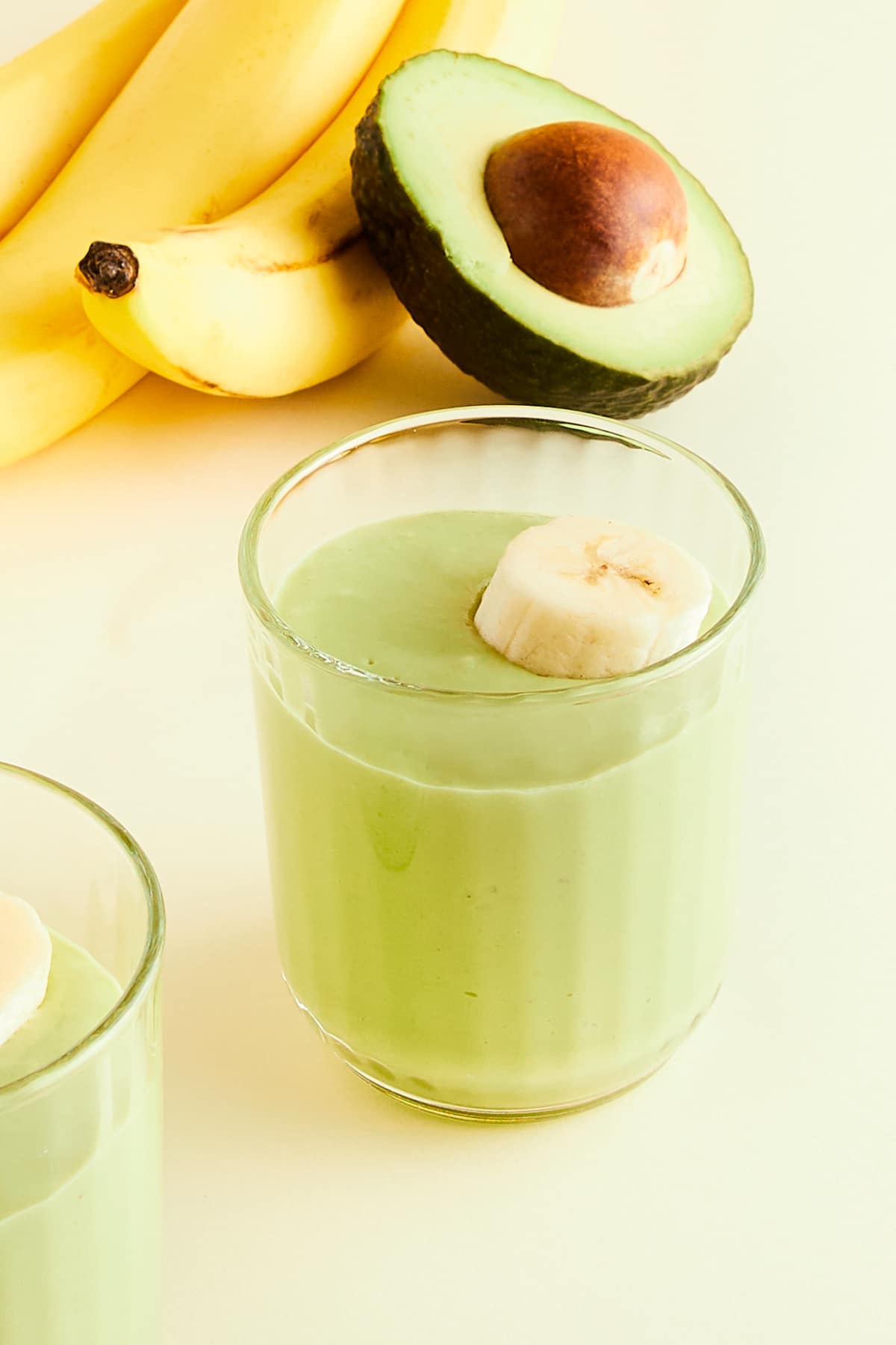 Two glasses of light green avocado smoothie garnished with banana slices. Whole bananas and a half avocado sit on the side.