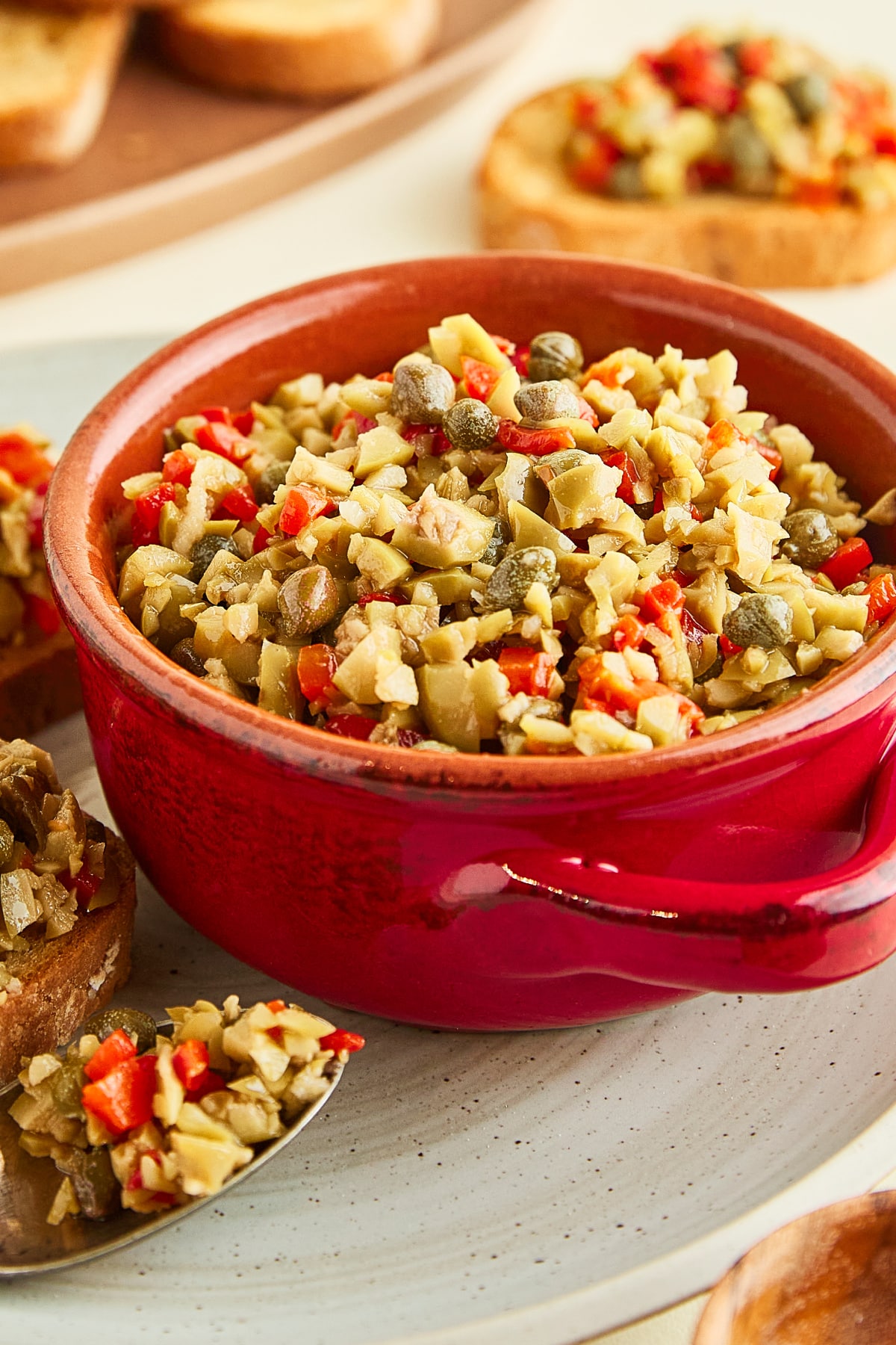 A red bowl of olive tapenade sitting on a rustic grey plate that holds two toasted baguette slices topped with tapenade, and a spoon filled with tapenade. Another plate of plain breads sits on the side.