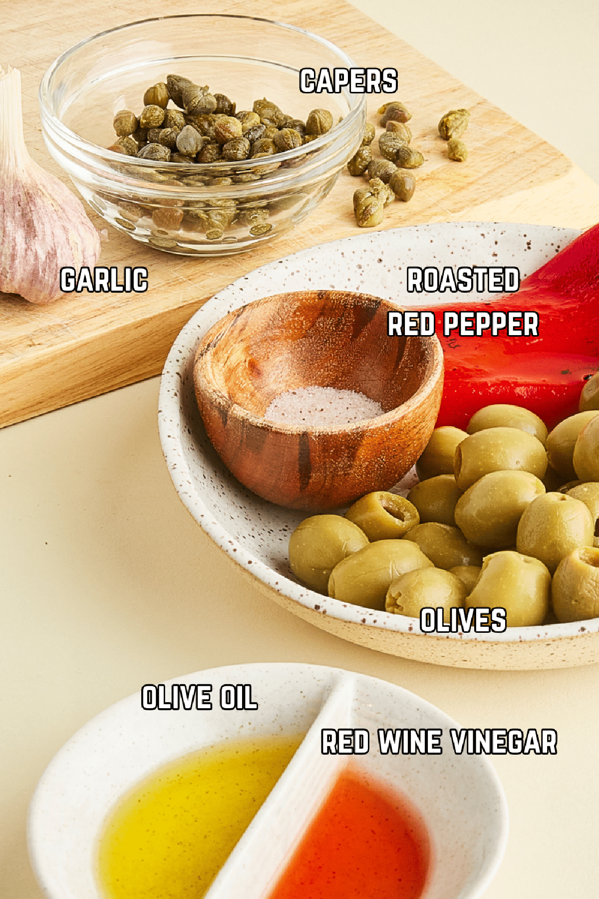 Ingredients to make tapenade sit in various bowls and boards: capers, garlic, roasted red pepper, olives, olive oil, and vinegar.