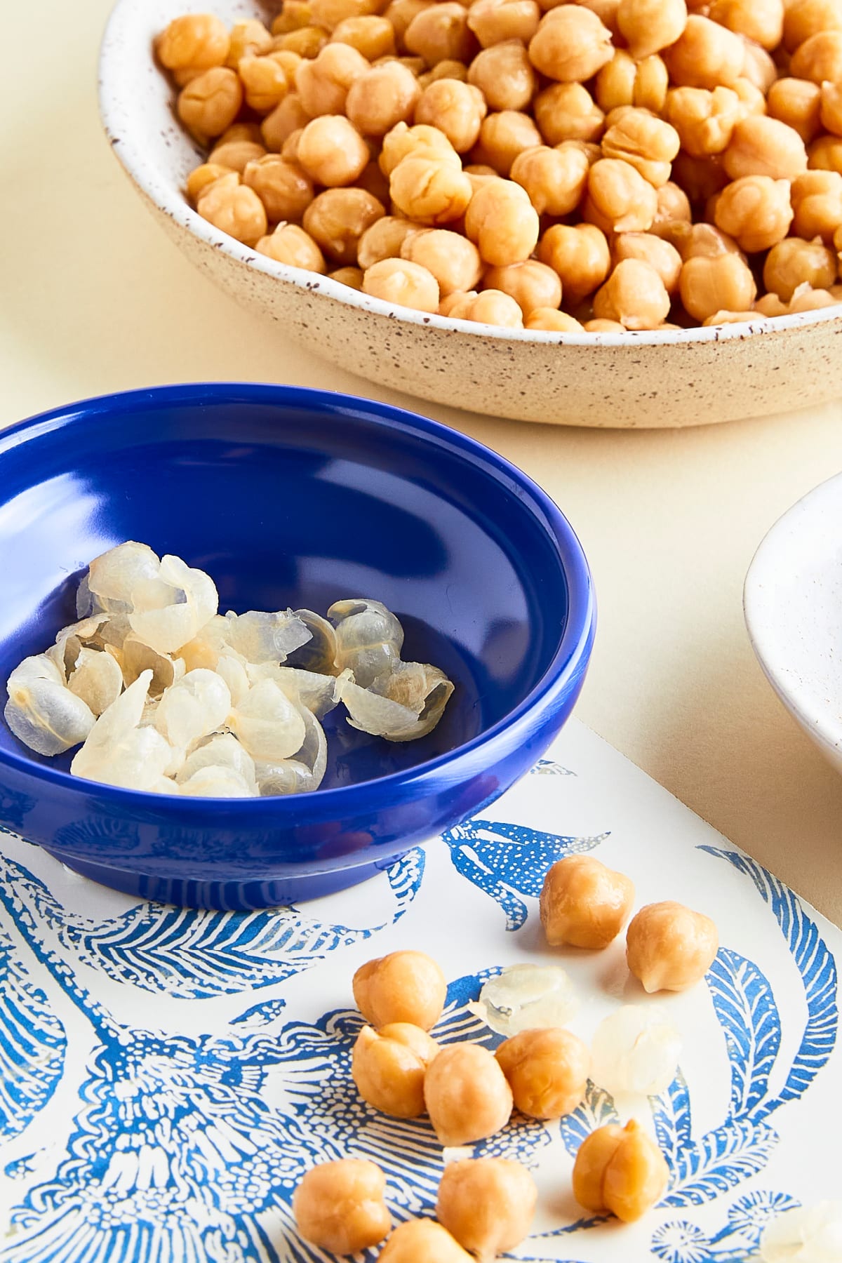 The process of peeling cooked chickpeas for a smoother hummus: a bowl of peeled chickpeas, another bowl of unpeeled chickpeas, and a small bowl of the peels.