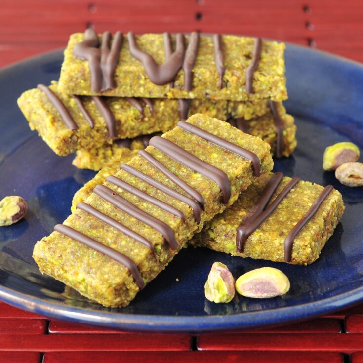 Light green pistachio lime bars drizzled with chocolate are stacked on a navy blue plate.