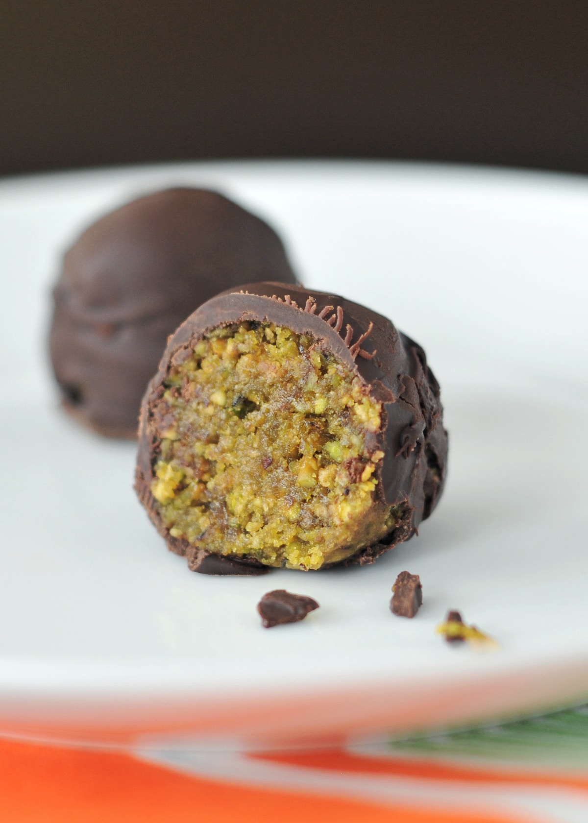 Two chocolate covered pistachio lime balls sit on a white plate; one ball has a slice out of it to show the light green inside.