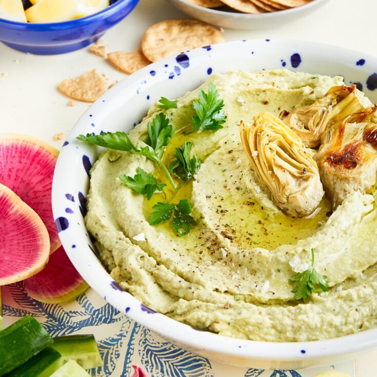 A serving bowl of roasted artichoke lemon hummus with brightly colored dipping veggies like cucumber, pink radish slices, purple and green endive, lemon wedges, and crackers.