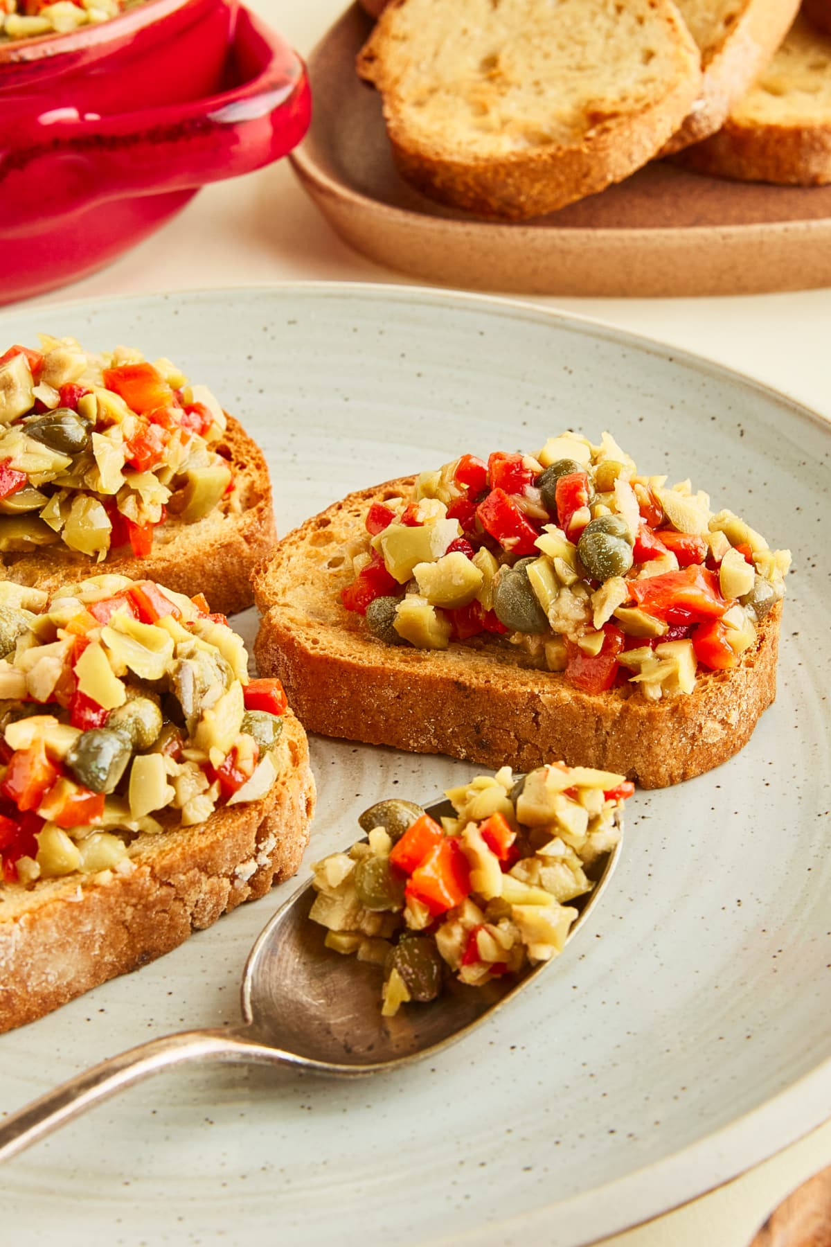 Toasted baguette slices topped with olive tapenade are arranged on a rustic grey serving plate. A spoon filled with tapenade sits on the plate, and a plate of plain toasted bread slices sits on the side.