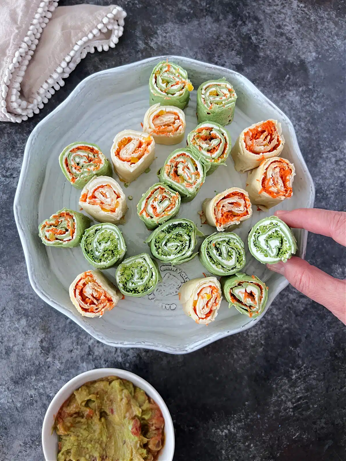 Colorful veggie pinwheels are stacked on a serving plate. Some pinwheels are made with green spinach tortillas, and some with plain white flour tortillas. All roll ups are filled with finely chopped veggies and dairy free cream cheese.