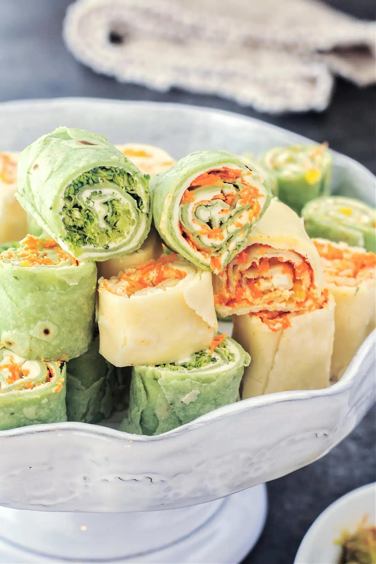 Colorful veggie pinwheels are stacked on a serving plate. Some pinwheels are made with green spinach tortillas, and some with plain white flour tortillas. All roll ups are filled with finely chopped veggies and dairy free cream cheese.