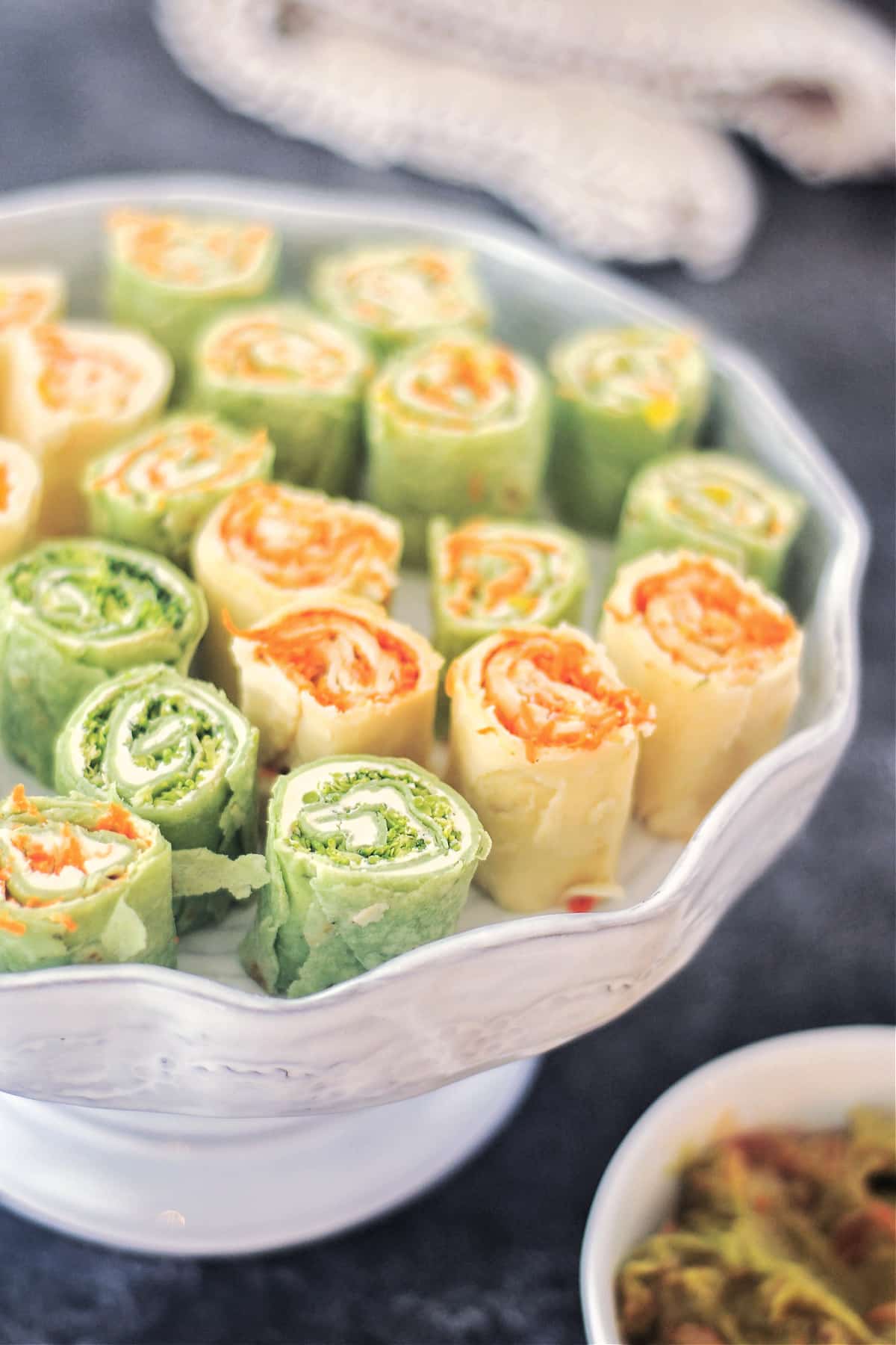 Colorful veggie pinwheels are stacked on a serving plate. Some pinwheels are made with green spinach tortillas, and some with plain white flour tortillas. All roll ups are filled with finely chopped veggies and dairy free cream cheese.