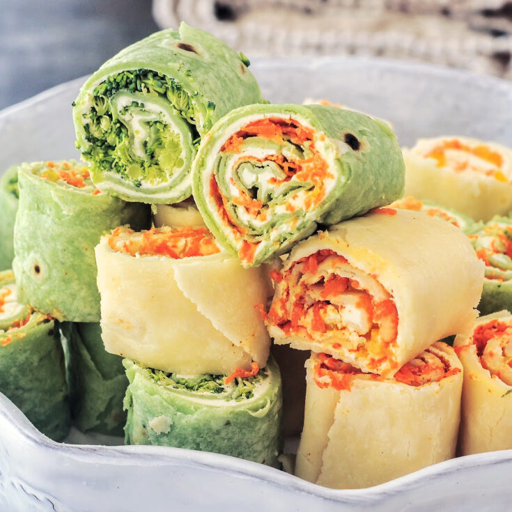 Colorful veggie pinwheels are stacked on a serving plate. Some pinwheels are made with green spinach tortillas, and some with plain white flour tortillas. All roll ups are filled with finely chopped veggies and dairy free cream cheese.