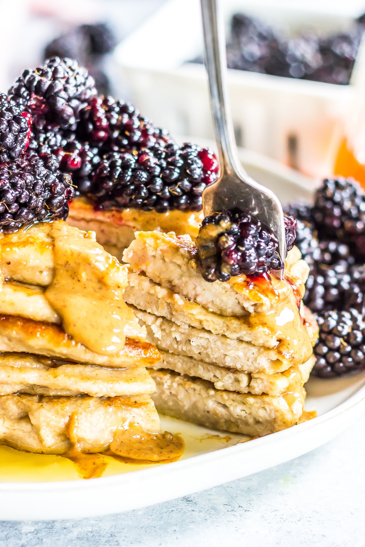 A stack of applesauce pancakes topped with peanut butter and blackberries has one bite sliced out with a fork stuck through the bite.