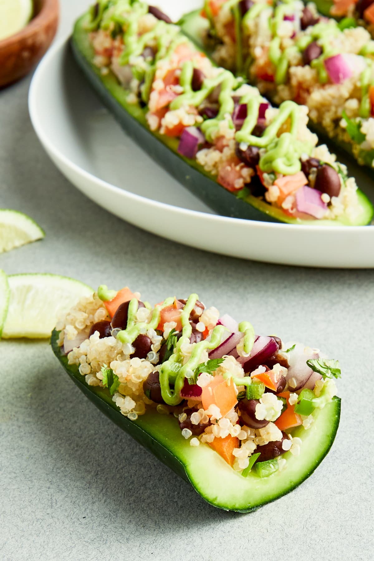 One half cucumber boat filled with a Mexican mixture of beans, quinoa, tomato and onion, and drizzled with a tangy avocado sauce sits on a grey surface in front of a serving plate of full tex mex cucumber boats.