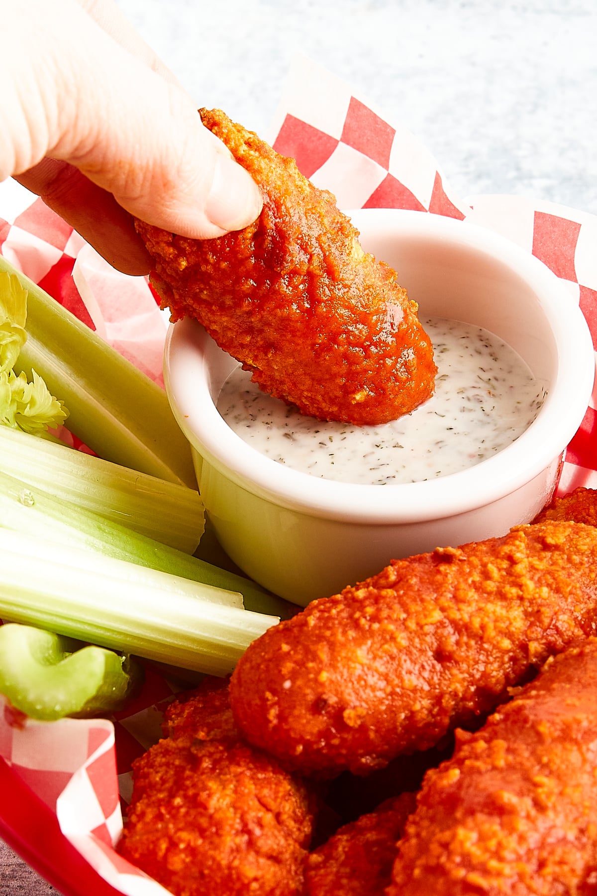 Soy free buffalo wings served in a red plastic basket lined with red and white checkered paper. Celery sticks and a small white bowl of ranch dressing are in the basket with the wings, and a hand is dipping one wing into the ranch.
