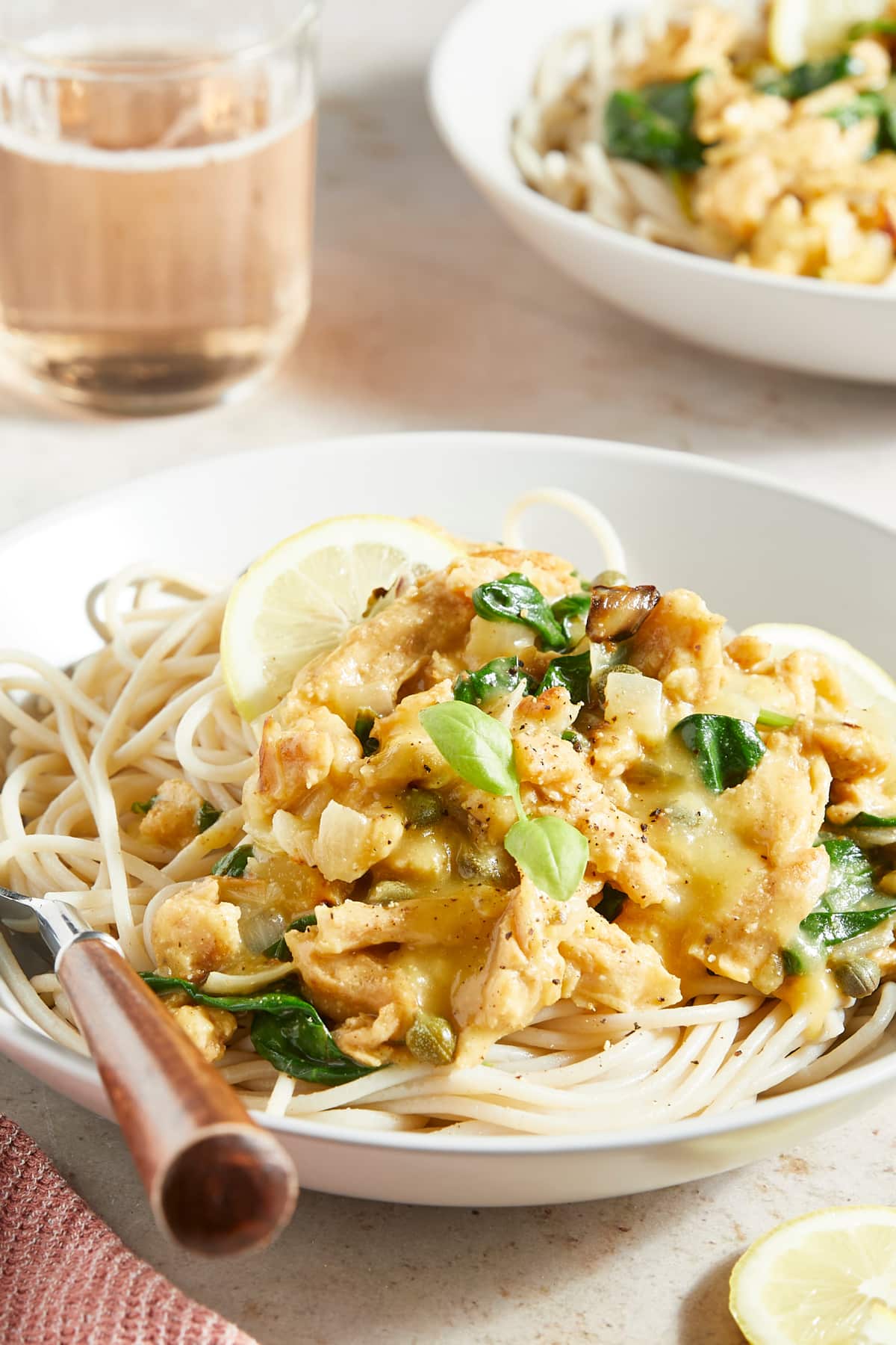 Side view of a plate of vegan chicken piccata over pasta noodles, garnished with lemon slices and fresh herbs. A wooden handled fork sits on the plate.