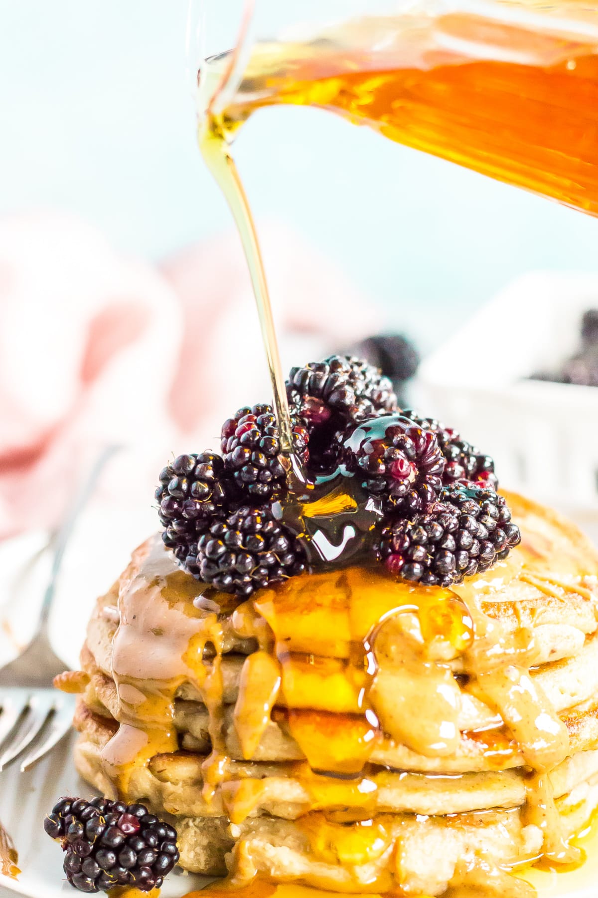 A small glass pitcher is pouring syrup over a stack of pancakes topped with peanut butter and blackberries.