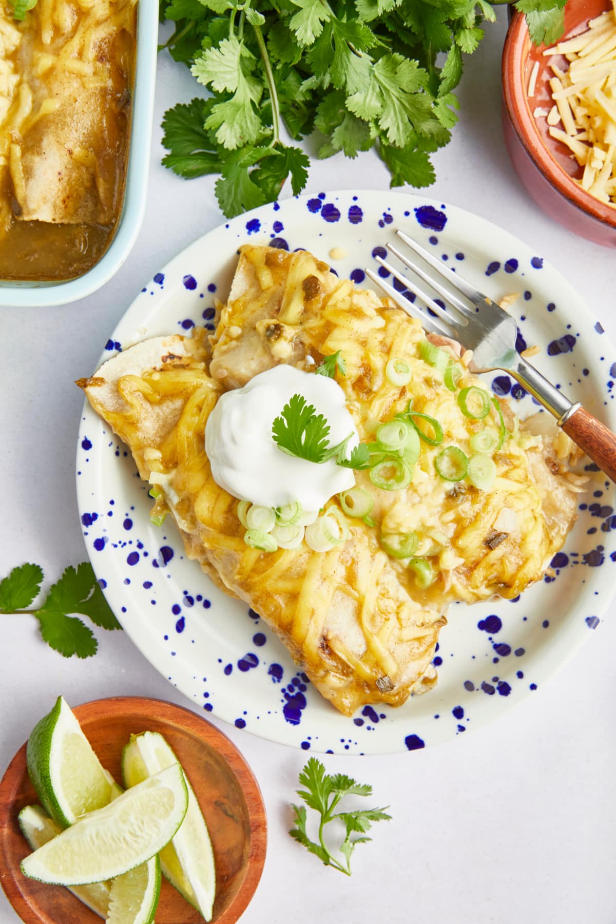 Overhead view of two dairy free enchiladas on a plate, garnished with sour cream, sliced green onions, and parsley. Sitting on the side is a bowl of lime wedges, a bowl of shredded cheese, more parsley, and the baking dish of enchiladas.
